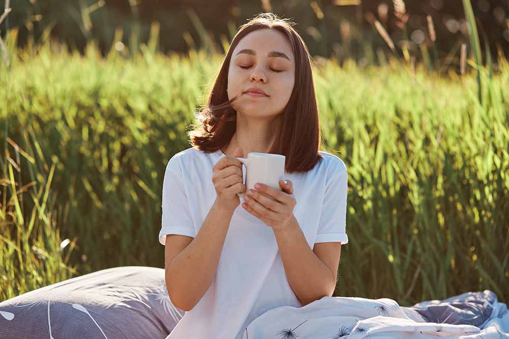 Zrelaxovaná tmavovlasá mladá žena v bílém tričku sedí venku v trávě a vychutnává si kratomový čaj